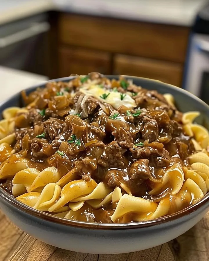 A hearty plate of French onion beef and noodles topped with melted cheese and herbs.